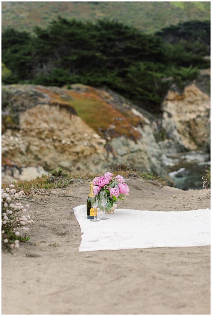 Glen Oaks Big Sur proposal picnic set up with champagne and pink peonies on a white blanket by film photographer AGS Photo Art
