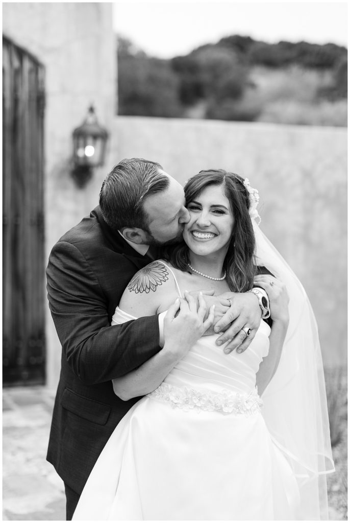 black and white Monterey wedding at The Club At Pasadera portrait of groom embracing his bride kissing her cheek while she smiles at the camera