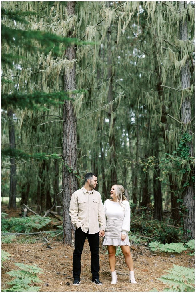 portrait of couple surrounded by trees as they hold hands and smile at each other