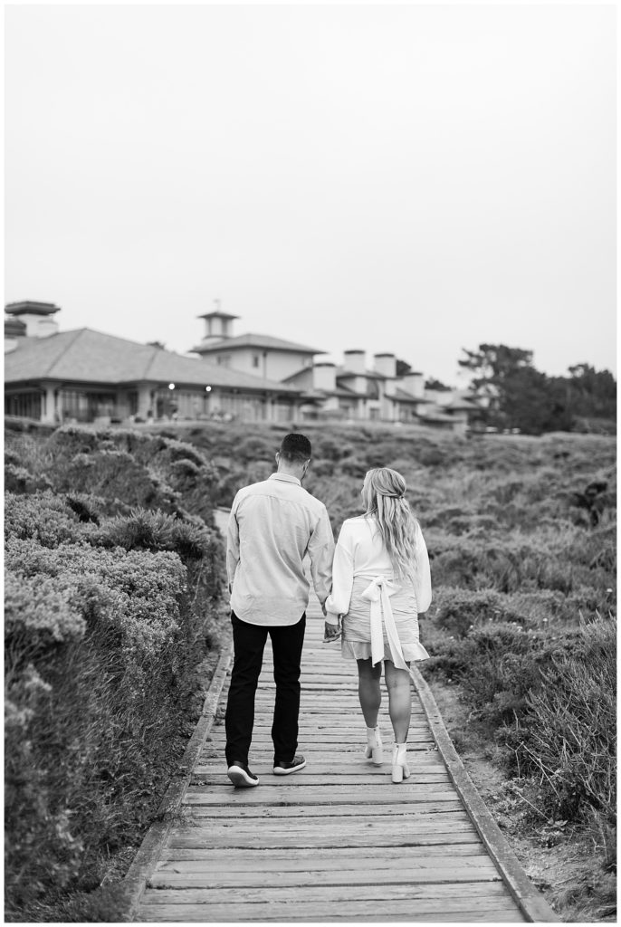 black and white Pebble Beach engagement session portrait of the couple walking hand in hand away from the camera down a wooden walkway