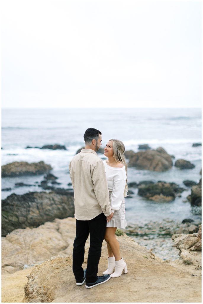 couple holding hands and smiling at each other on the coastline with waves behind them