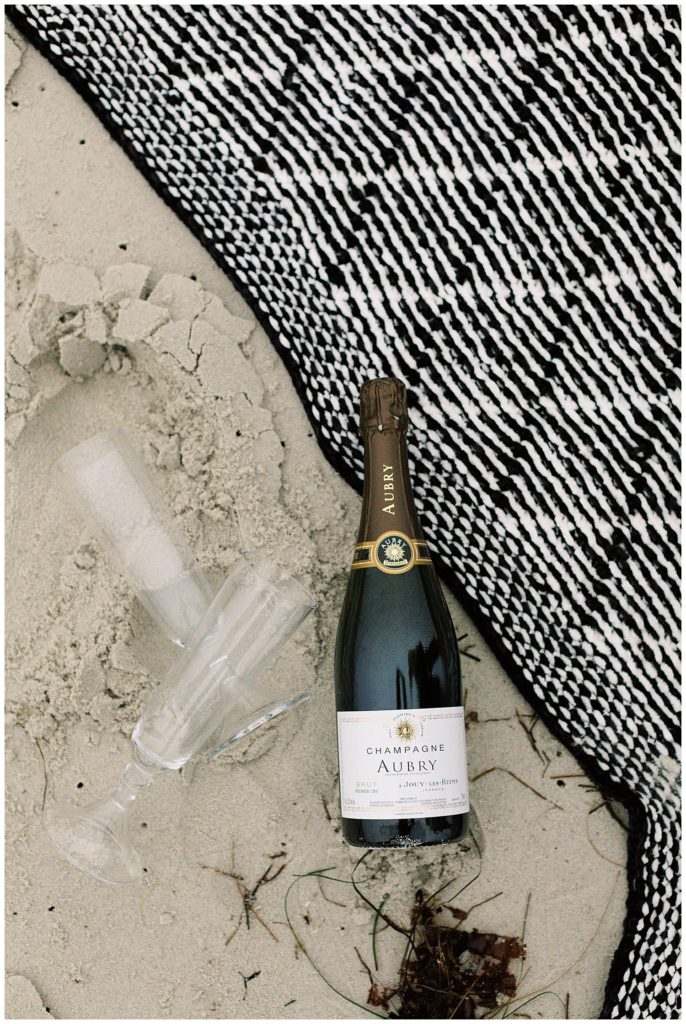 bottle of Aubry champagne and two empty glass flutes on the sand near a black and white picnic blanket