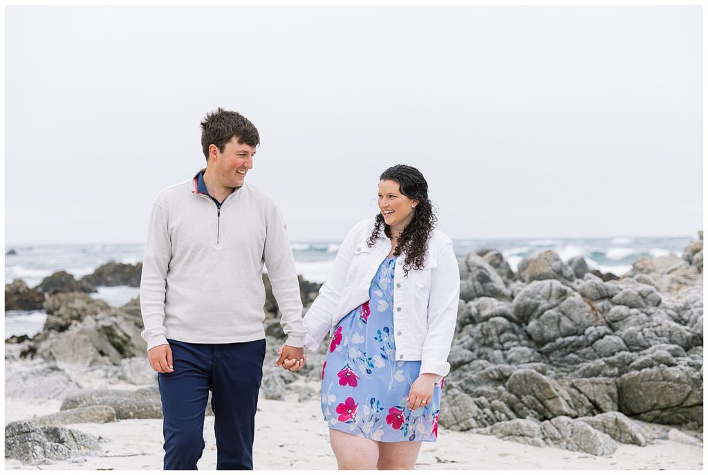 couple smiling and walking hand in hand with the beach and rocks behind them
