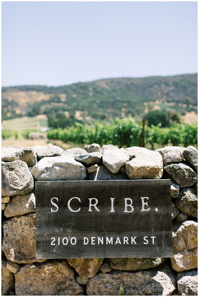 wooden sign on a stone wall reading "SCRIBE 2100 DENMARK ST" with a view of winery vineyards and green hills in the background