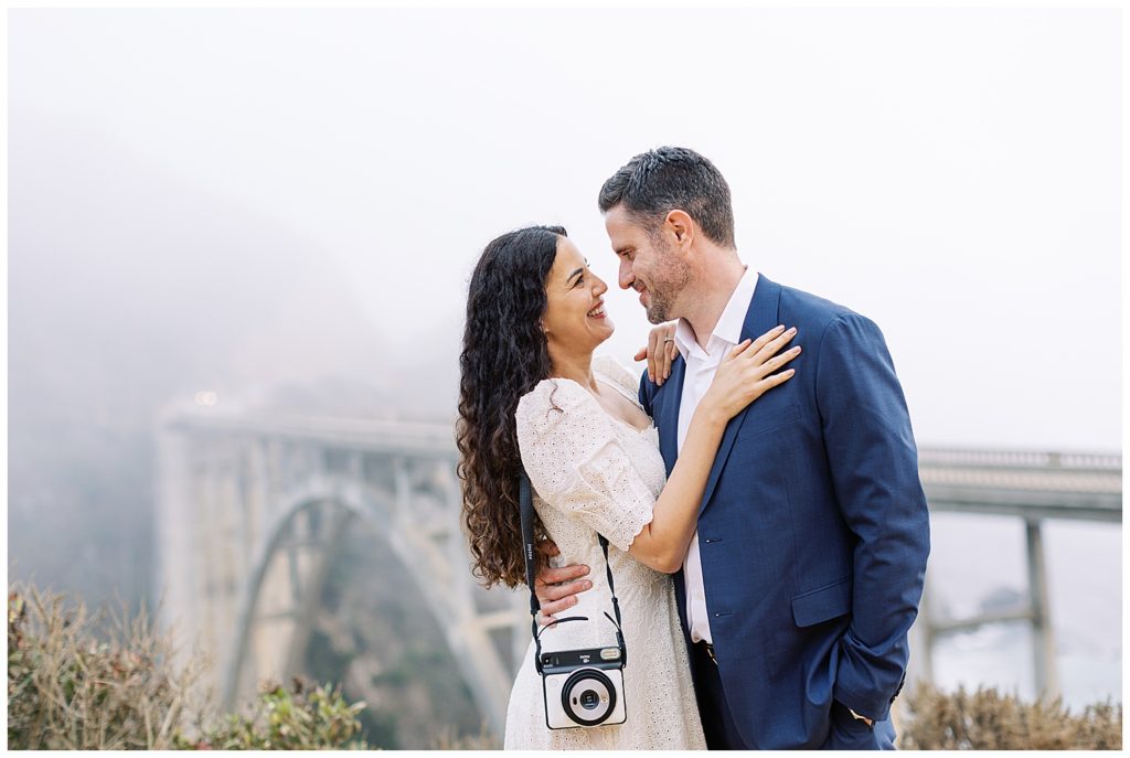 couple embracing and smiling at each other with Bixby Bridge behind them