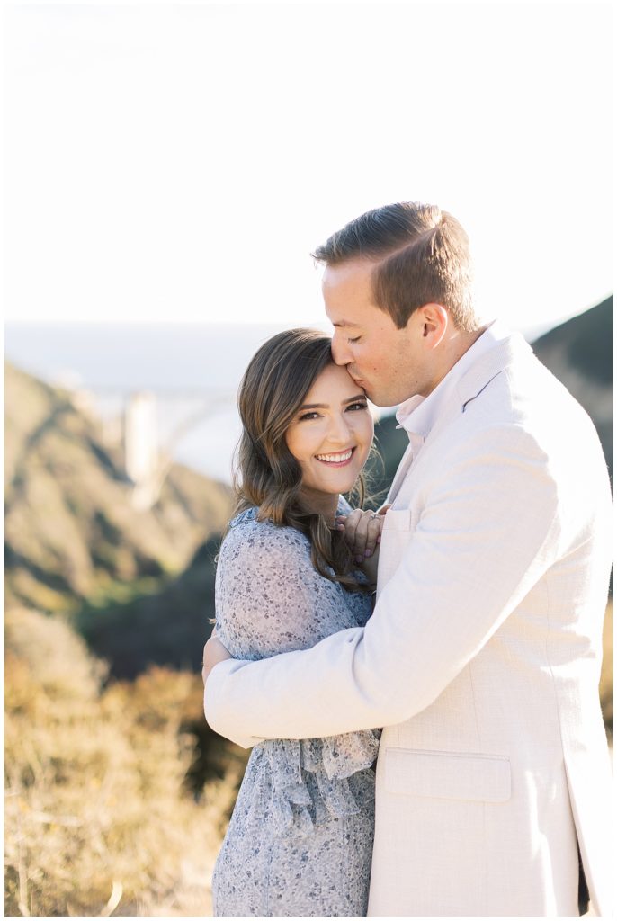 groom kissing his new bride on the forehead while she smiles at the camera with Bixby Bridge behind them both