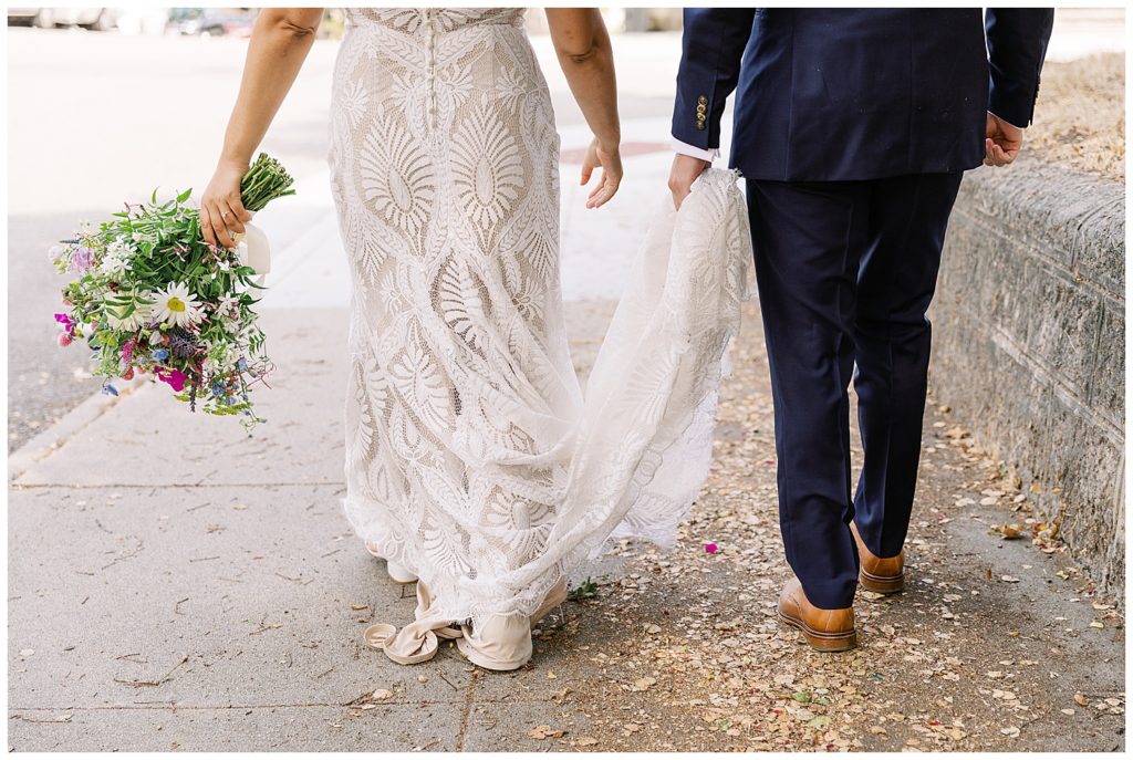 couple from the waist down walking away from the camera with the groom holding the train of his bride's gown
