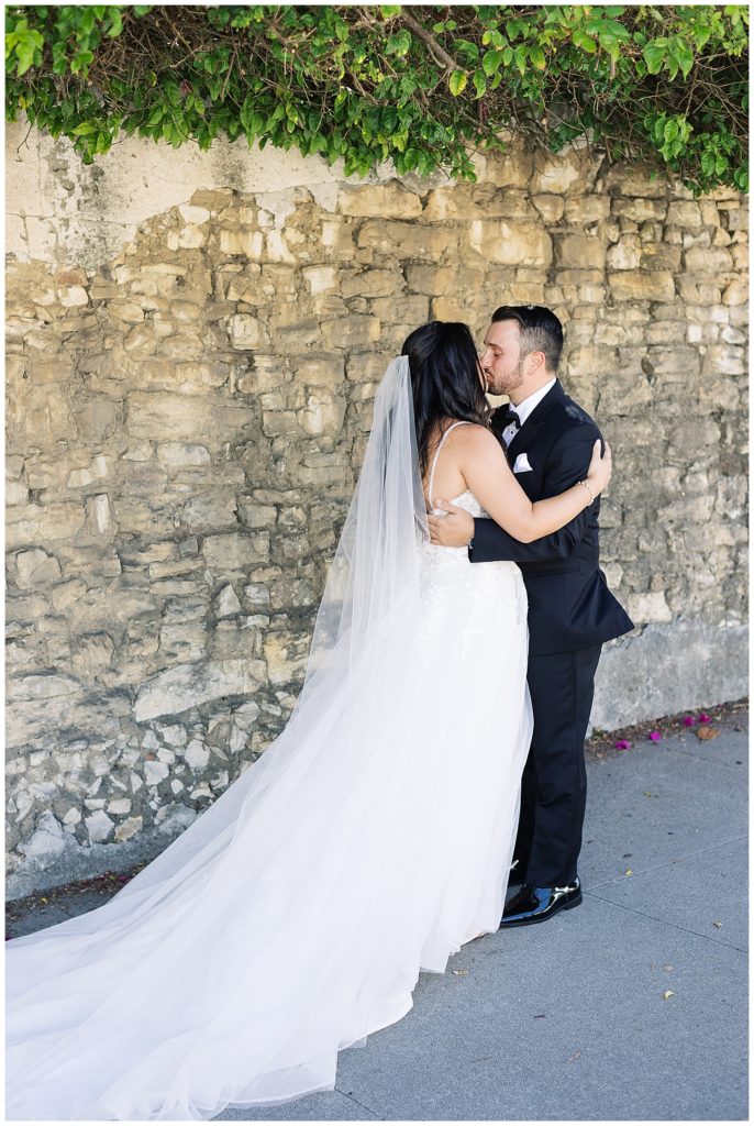 couple sharing a kiss in front of a stone wall with green leaves spilling out over top at their Classic Monterey Bay Wedding