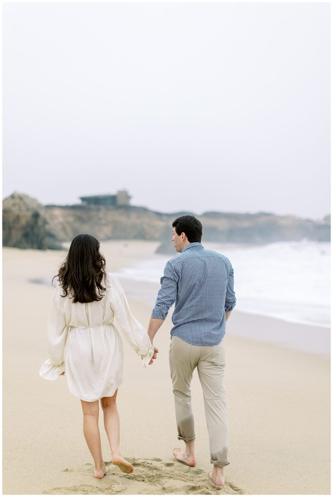couple hand in hand and walking away from the camera on the sand during their engagement session in Big Sur by film photographer AGS Photo Art