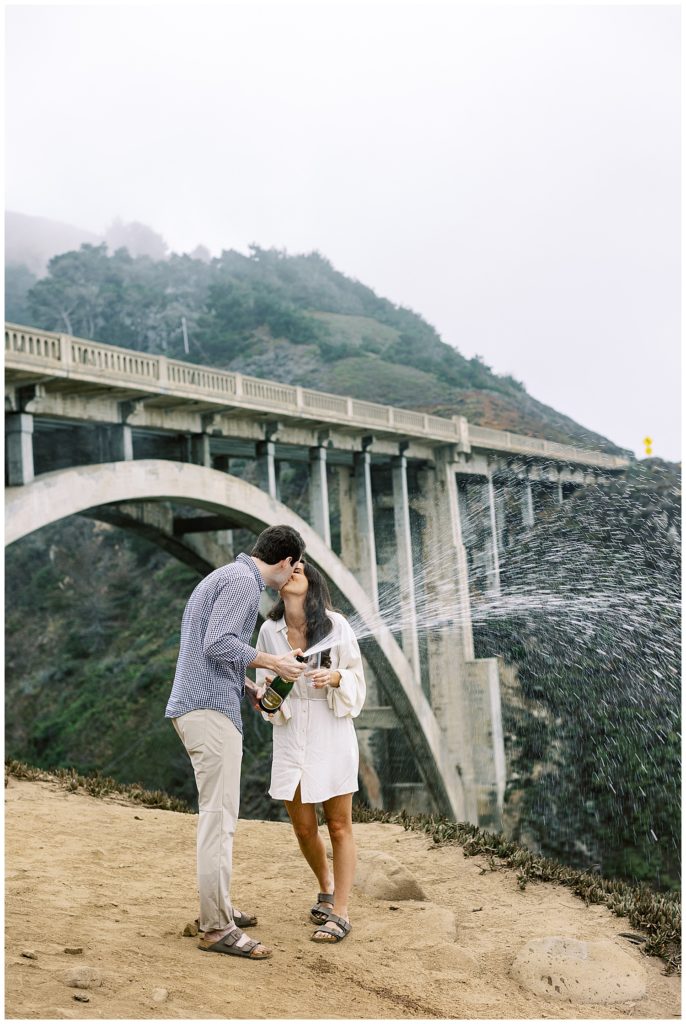 portrait of the couple sharing a kiss and popping champagne with Bixby Bridge in the background of their Big Sur engagement shoot