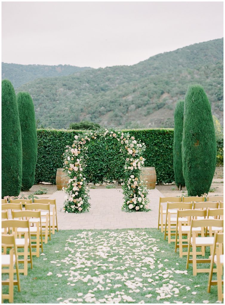 ceremony archway made of white roses with white petals scattered on the grass leading up to that