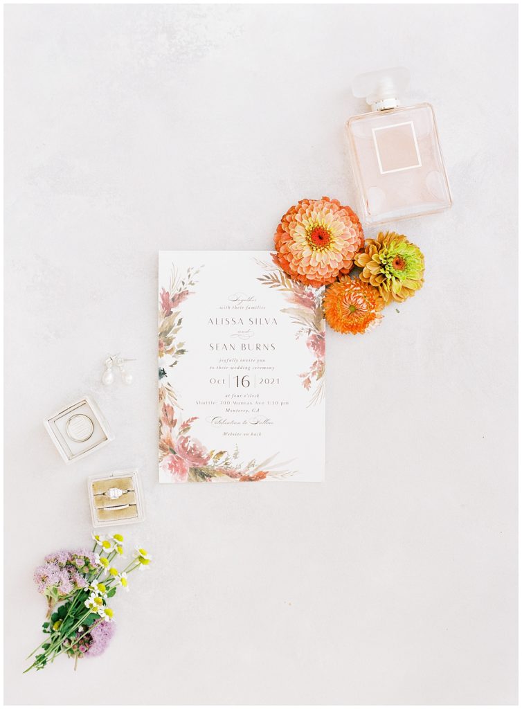 Wind & Sea Estate Big Sur wedding flat lay of bridal details including the invitation suite, florals by Big Sur Flowers, a pink glass perfume bottle, pearl earrings, and a family heirloom wedding ring