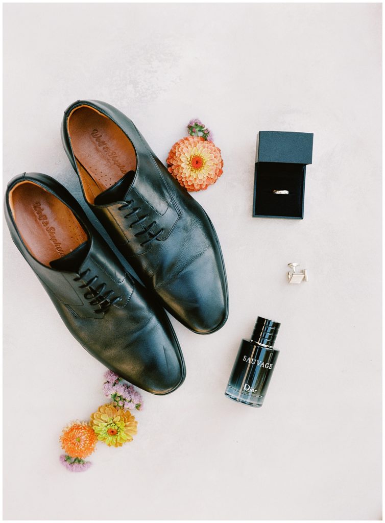flat lay of the groom's wedding shoes by Wolf & Shepherd, florals by Big Sir Flowers, Sauvage cologne by Dior, and rings by Derco Diamonds