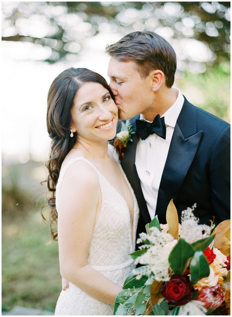 bride smiling at the camera and holding her bouquet by Big Sur Flowers while her groom kisses her cheek