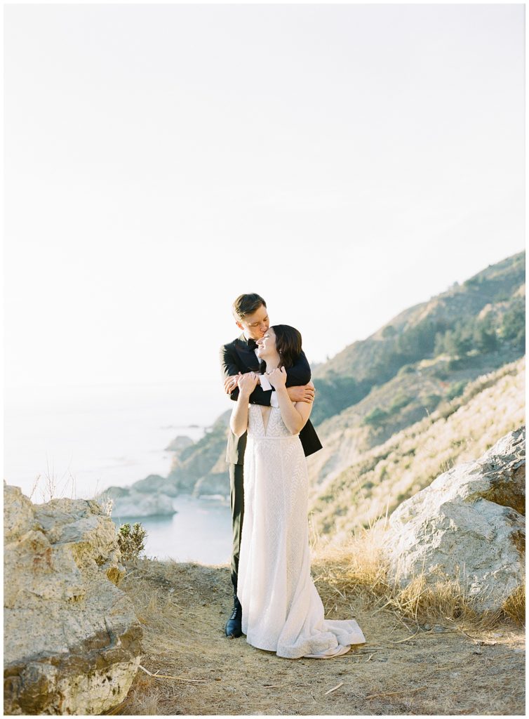 portrait of the groom embracing his bride from behind and kissing her forehead with the view of Big Sur behind them by film photographer AGS Photo Art