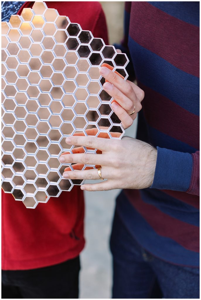 close up of couple's hands with engagement rings on them as they hold a honeycomb mirror