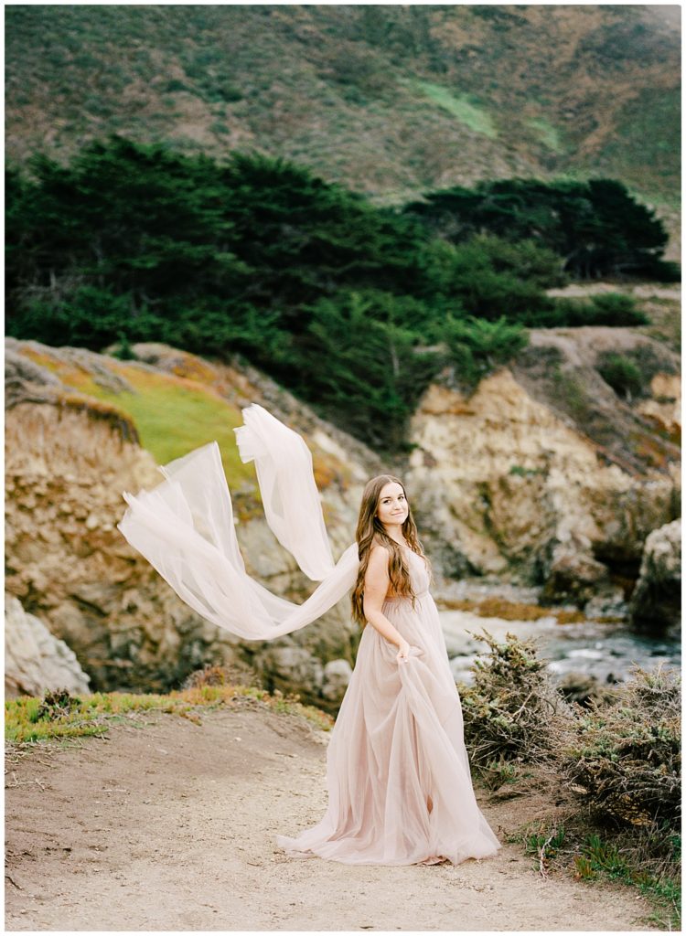 portrait of a bride wearing a pale blush dress whose sleeve sashes flow in the wind in Big Sur by film photographer AGS Photo Art