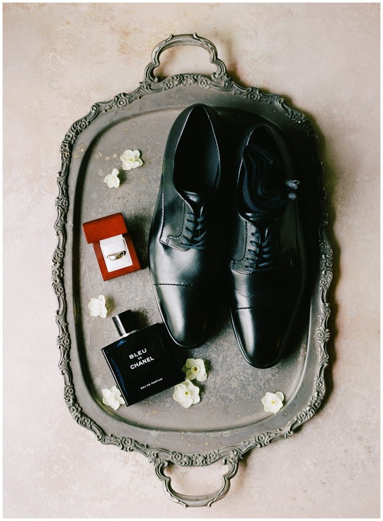 groom's wedding details on a silver platter featuring his shoes, ring, cologne, and flowers
