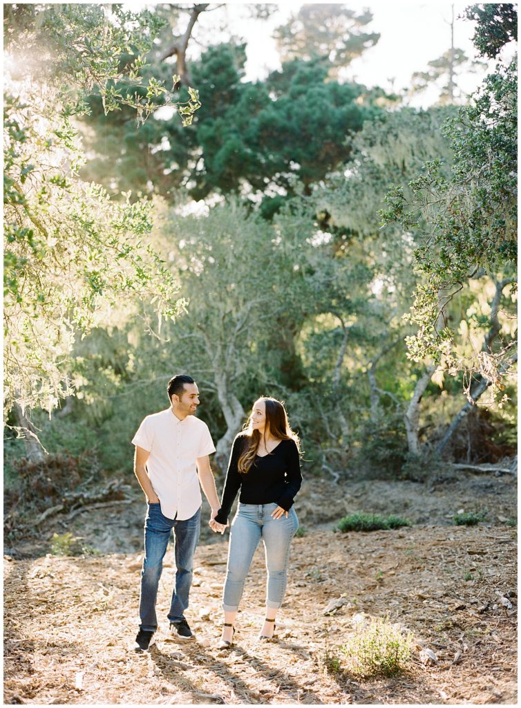 Pebble Beach engagement session in the forest with the couple walking hand in hand smiling at each other