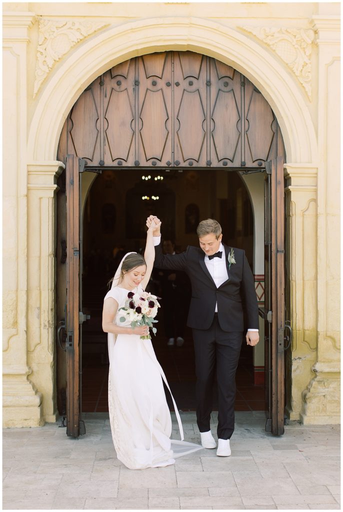 bride and groom walking out of San Carlos Cathedral with their hands together in the air