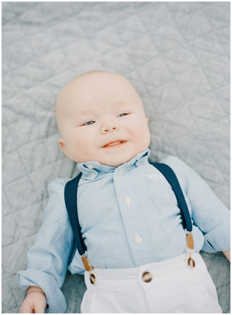 portrait of sixth-month Wills smiling up at the camera while he lies down on a blue quilt