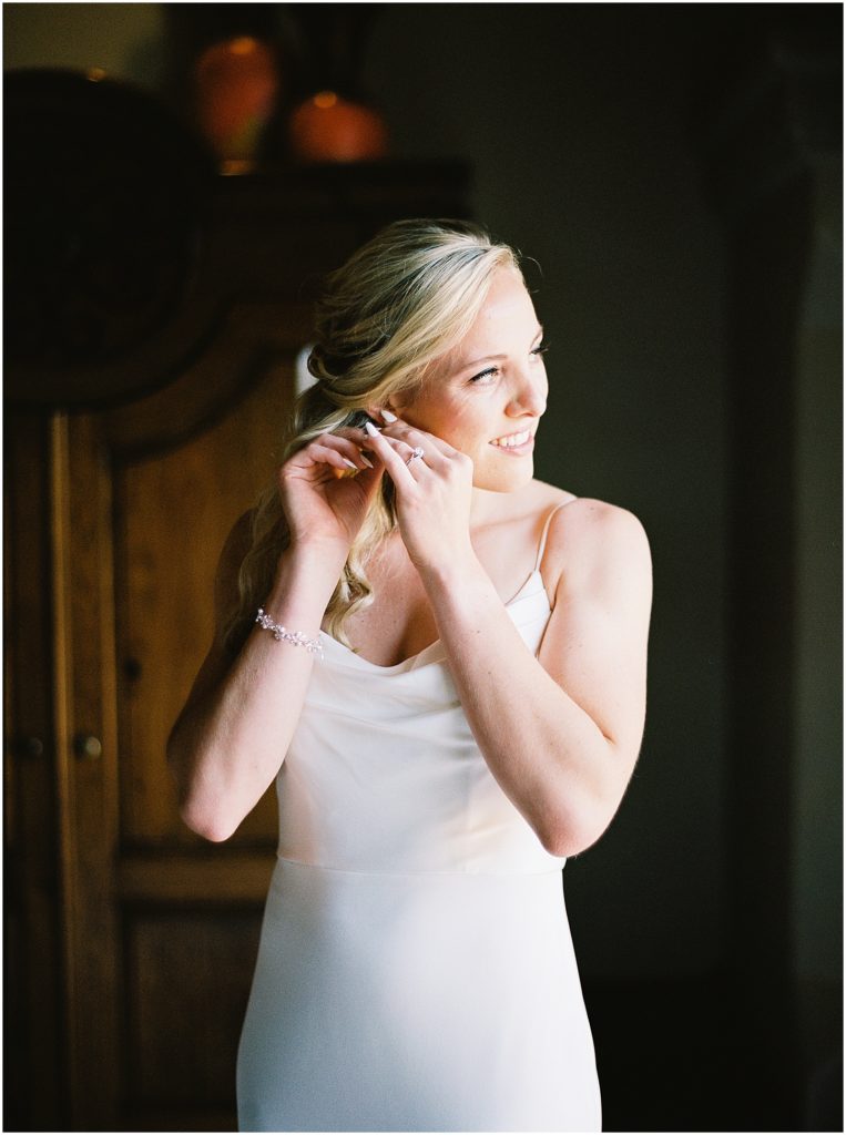 portrait of bride standing by window fixing jewelry by film photographer AGS Photo Art