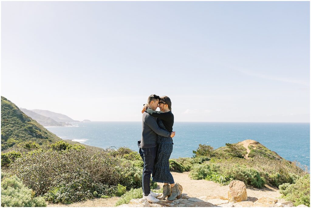 photo of newly engaged couple embracing in a hug with the big Sur coastline behind them, at a surprise proposal in Big Sur