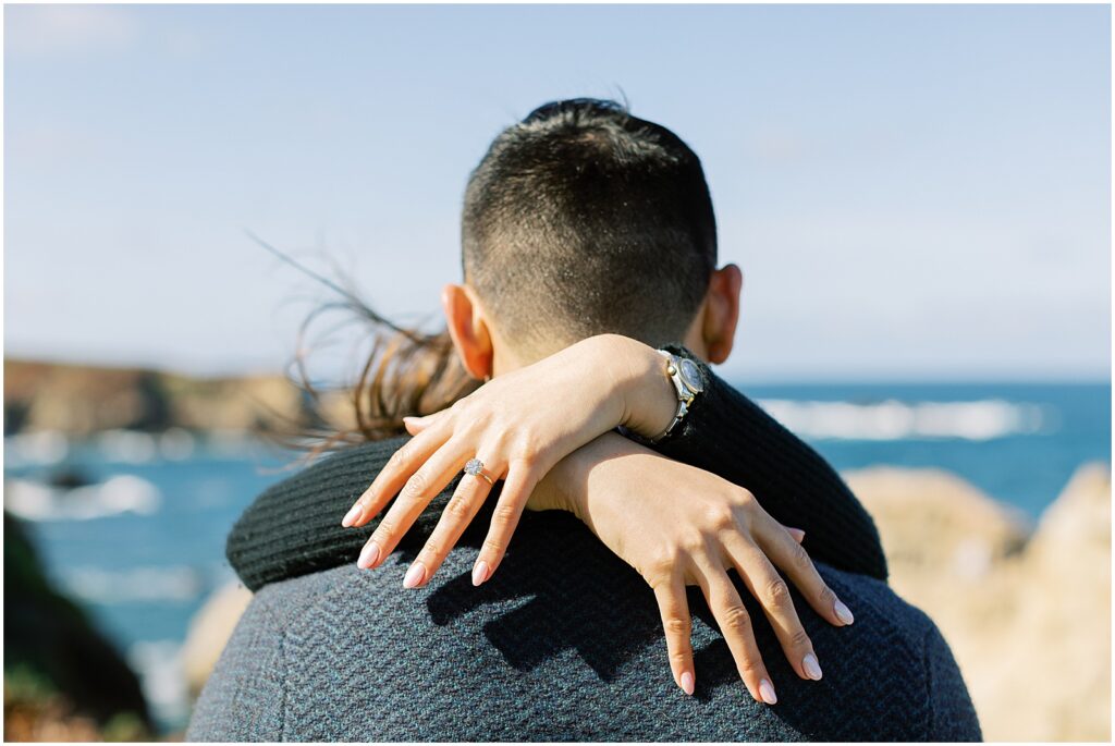 photo of an engagement ring, close up of woman's hands draped over her fiancee's shoulders at a surprise proposal in Big Sur