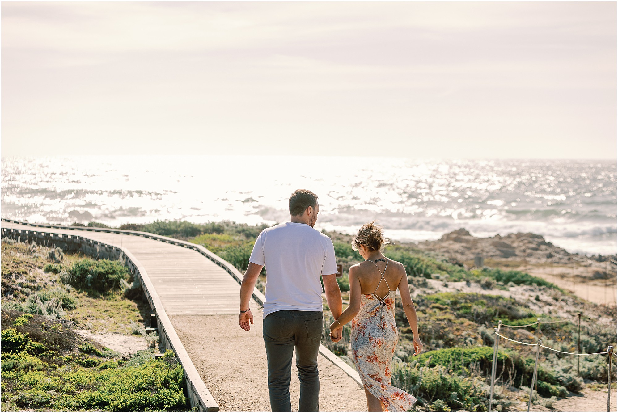 Asilomar Beach Proposal Photographer captures Josh and Tasha walking hand in hand down the Pacific Grove boardwalk, sunlight sparkling on the ocean.