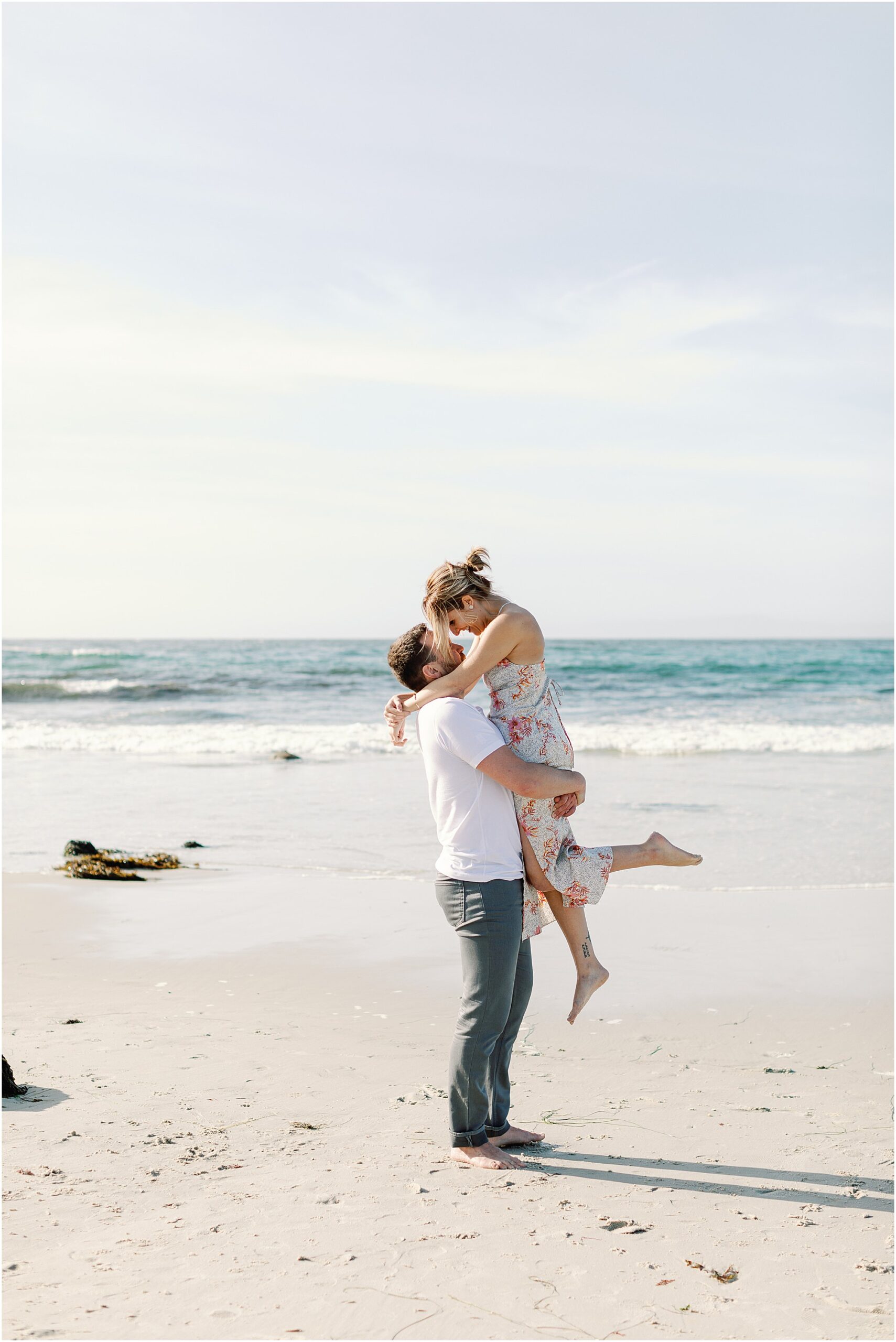 Josh lifts Tasha joyfully on the sand as waves roll in, captured by Asilomar Beach Proposal Photographer in Pacific Grove.
