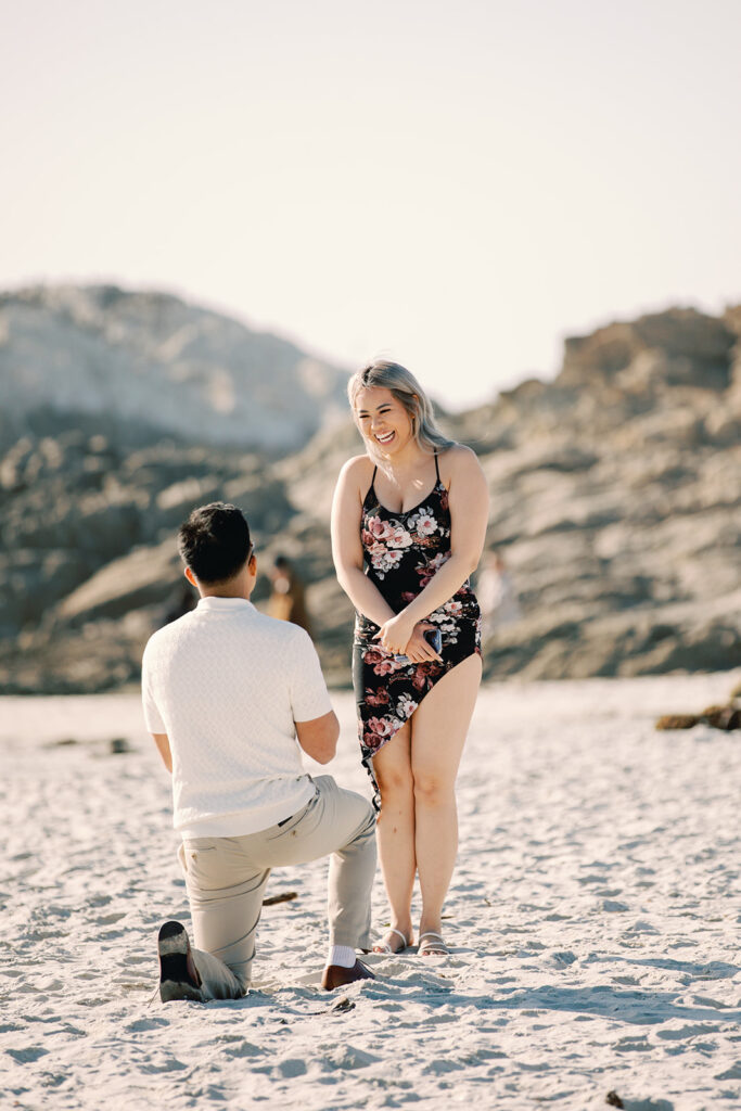 Mark on one knee with his back to the camera as Vivian smiles brightly in a black floral dress during their surprise Pebble Beach proposal on the sand with rocks behind them.