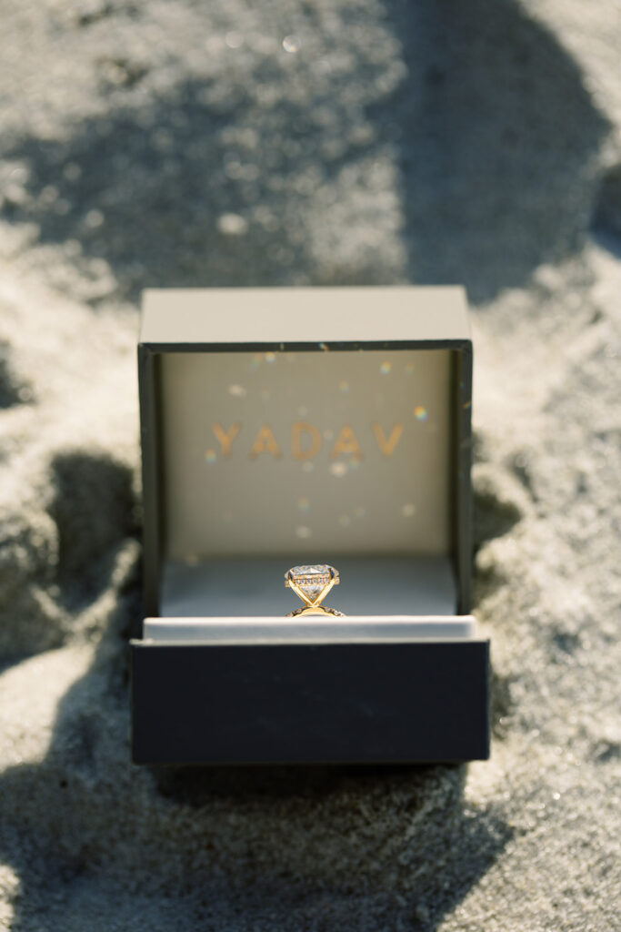 Close-up of sparkling round engagement ring in a brown Yadav box, reflecting sunlight while sitting in the sand during Pebble Beach proposal.