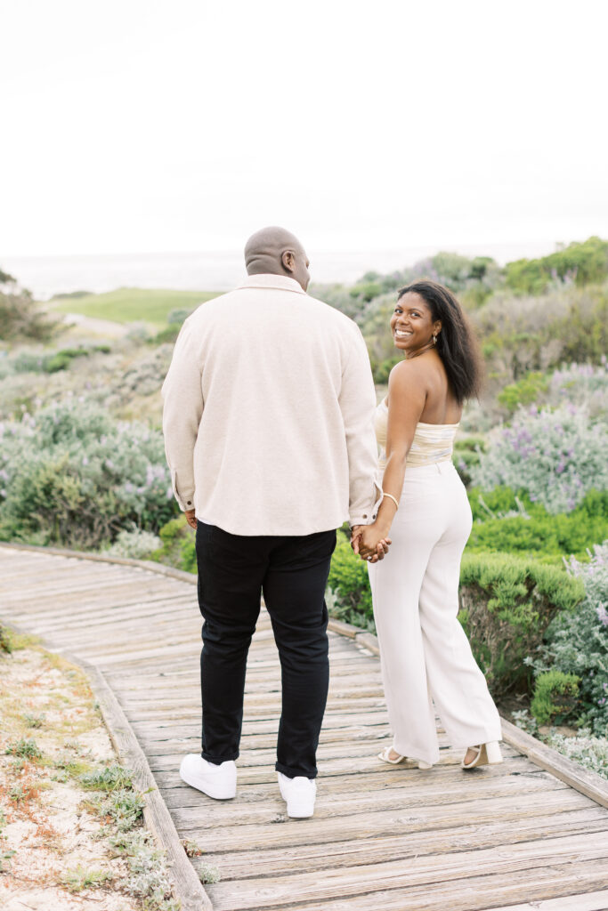 Couple walking across Spanish Bay beach with the grassy dunes behind them.