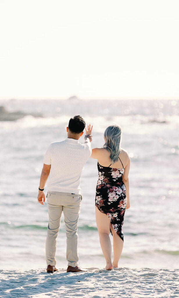 Mark and Vivian facing the ocean at Spanish Bay, holding up her engagement ring after saying yes, captured by Pebble Beach proposal photographer.