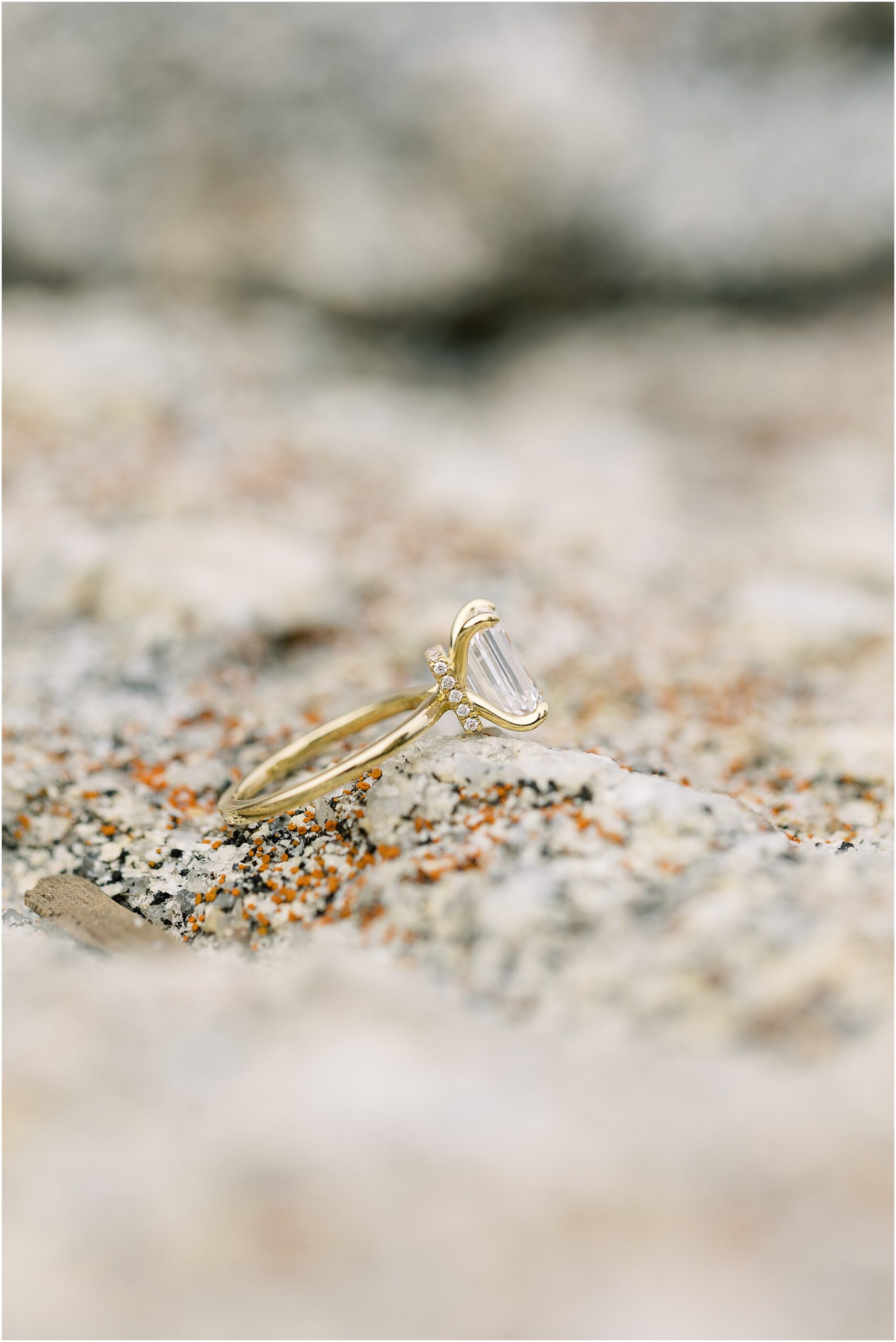 Emerald cut engagement ring partially nestled in the sand with the ocean softly blurred in the background.