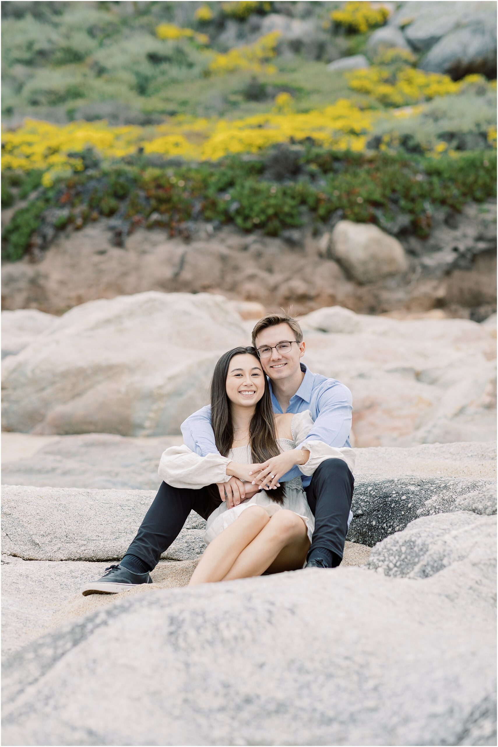 Couple wrapped in an embrace with the rocky coast and flowers behind them.