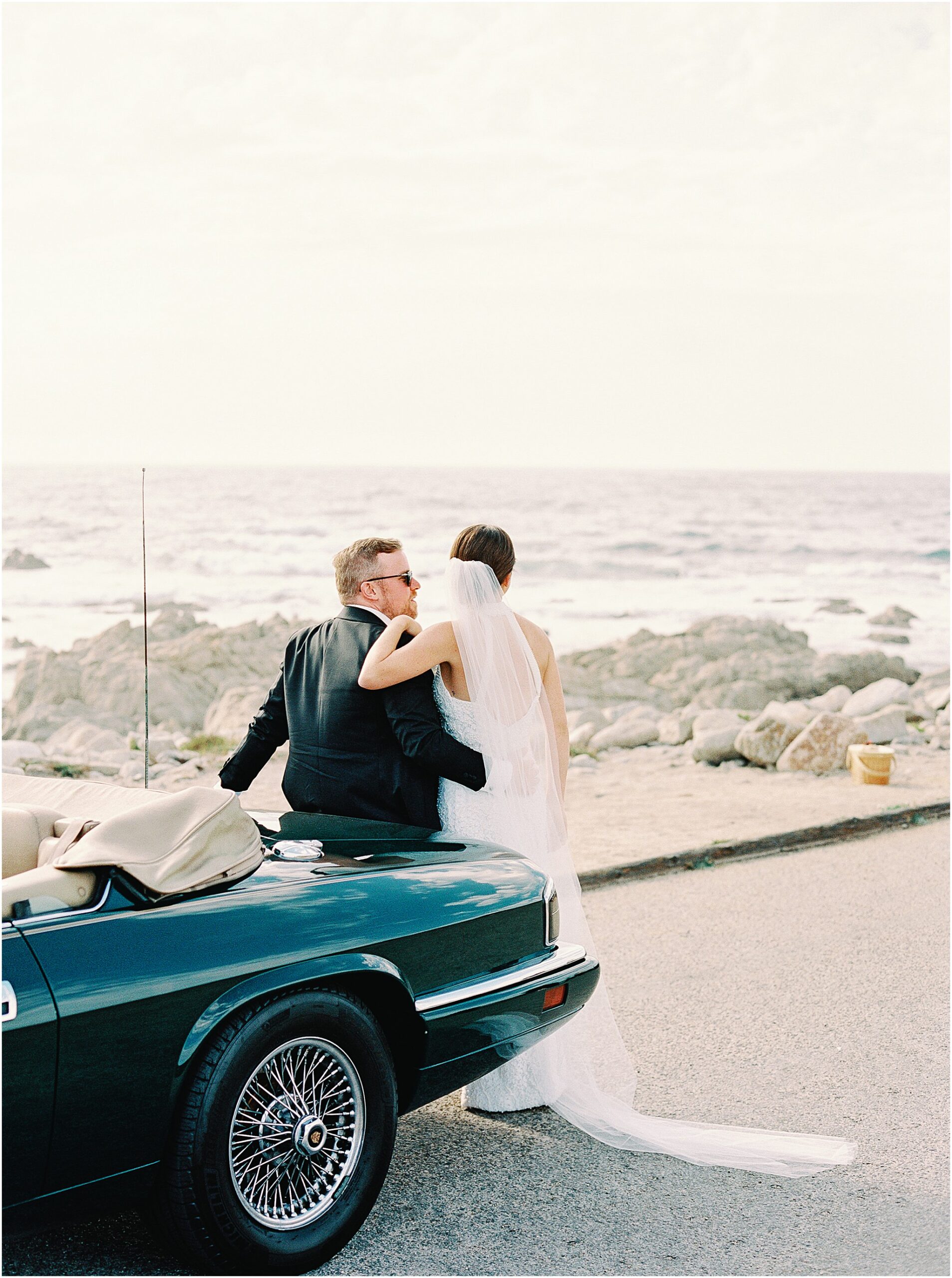 Couple sitting on vintage Jaguar admiring coastal view.
