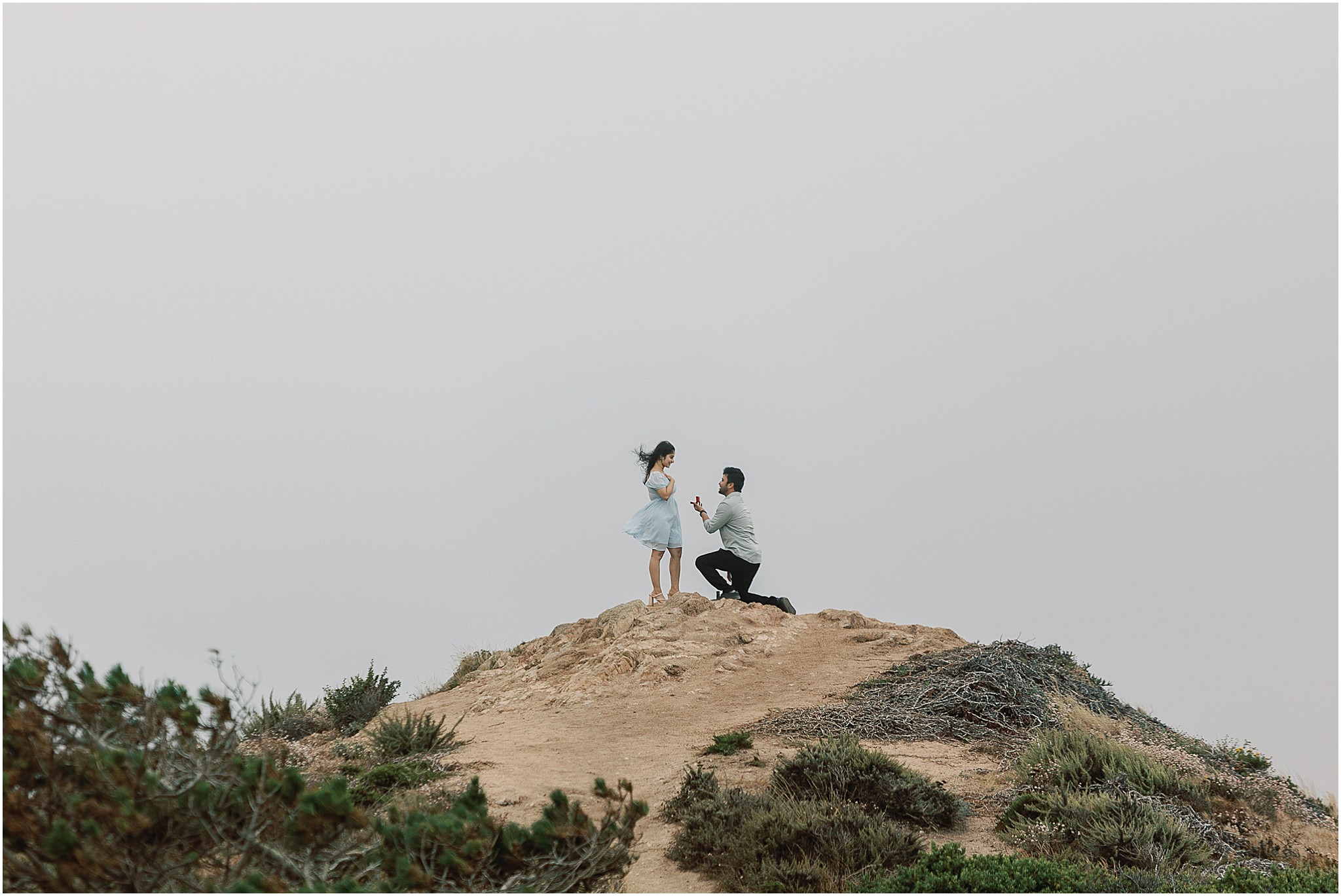 Groom on one knee proposing on the beach in Big Sur as she reacts with surprise and joy.