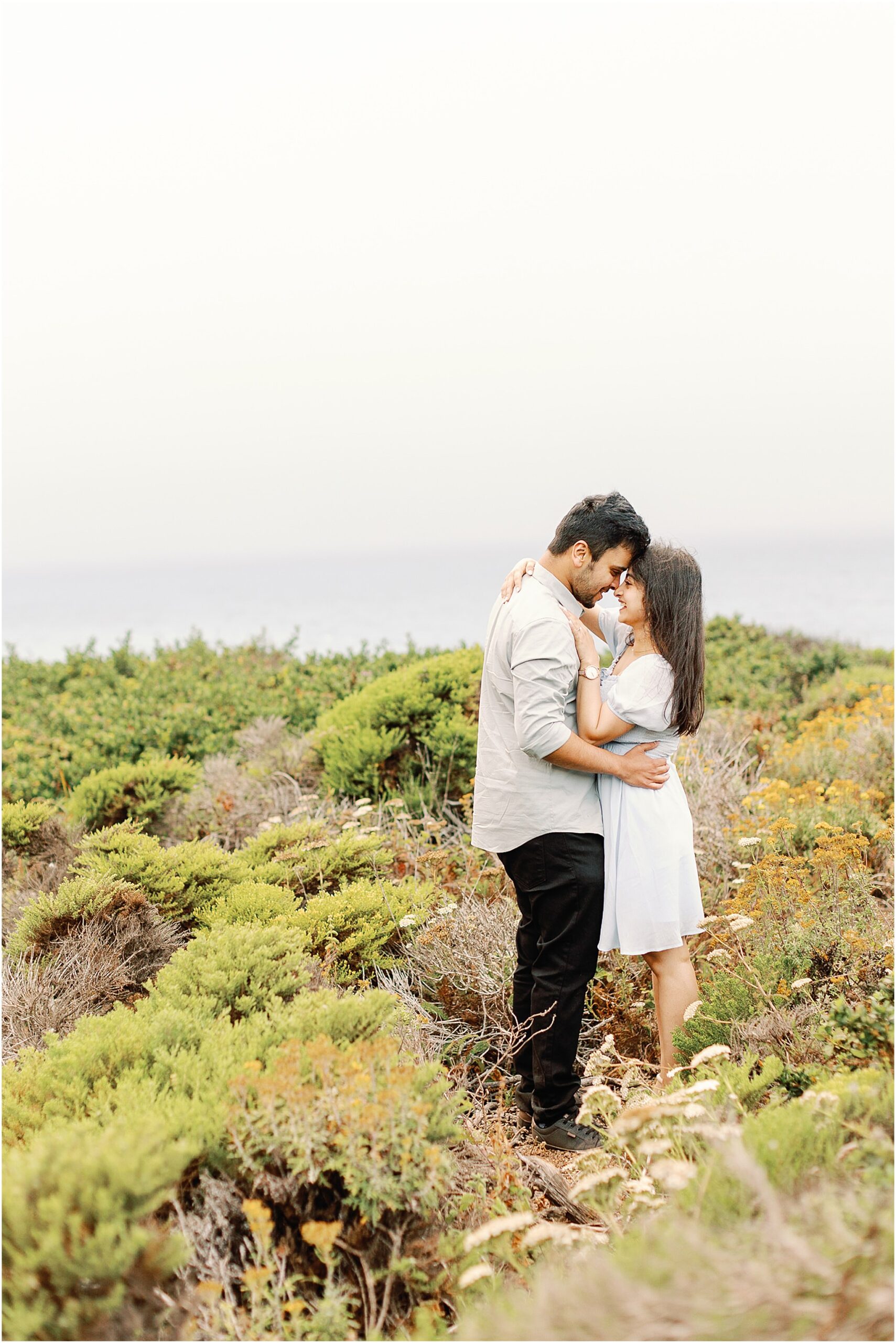 The couple shares a warm embrace after the proposal with misty ocean views behind them.