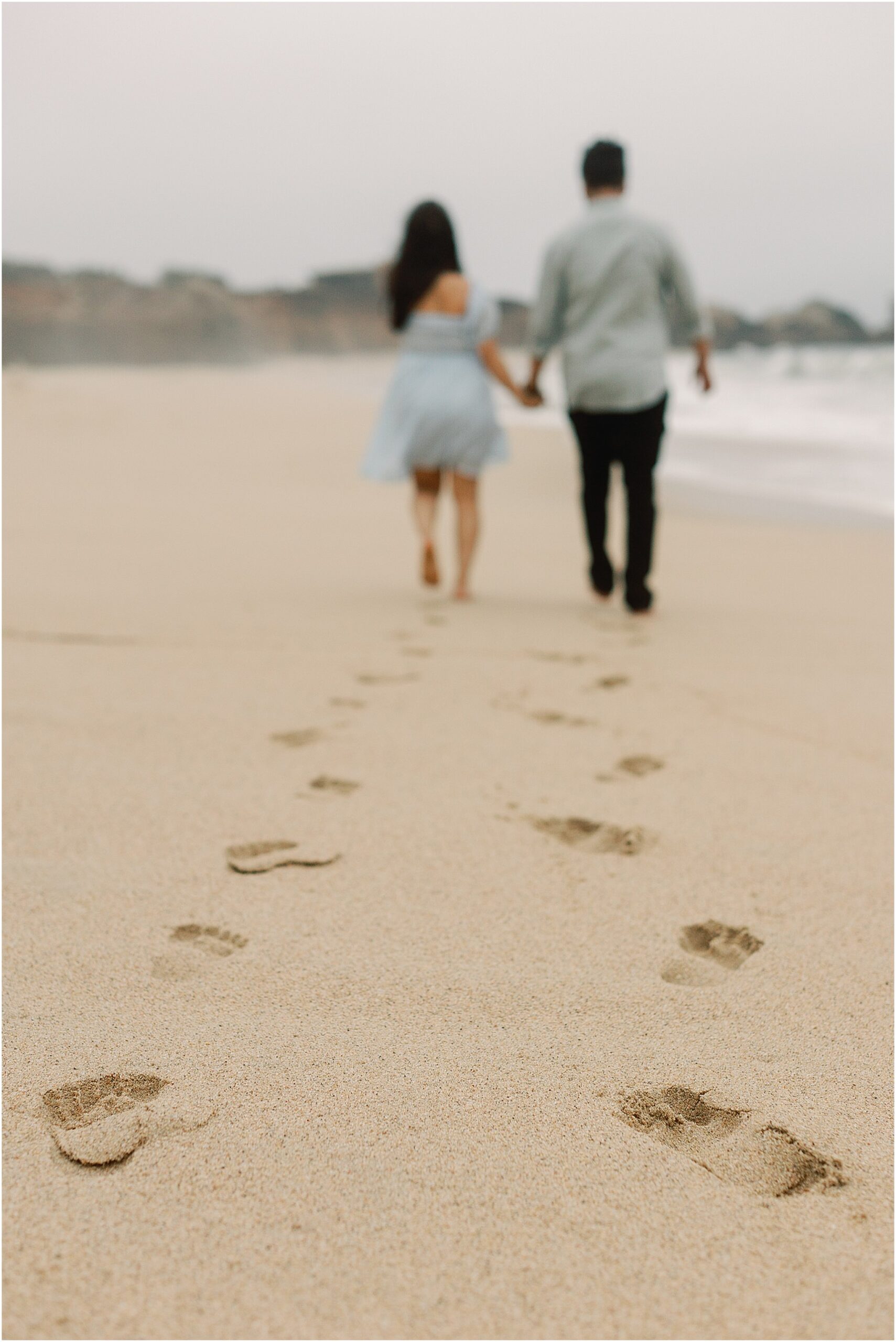 Couple holding hands and walking along the waterline during a romantic proposal session.