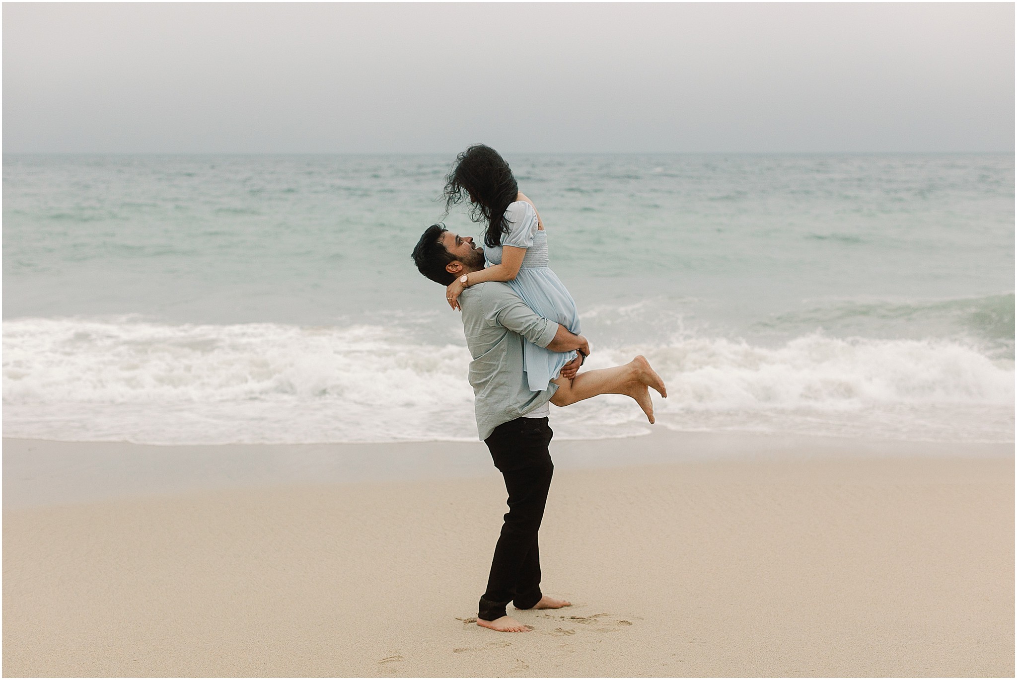 Groom lifts bride to be in a playful embrace after the proposal on a misty Big Sur beach.