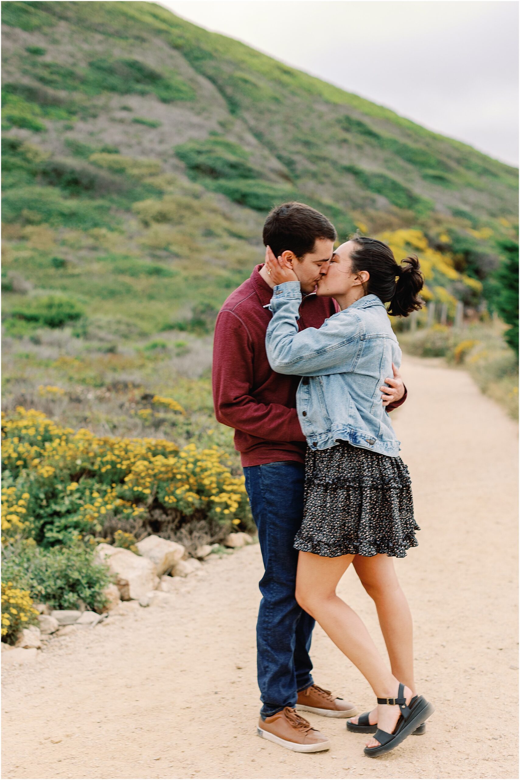 Couple kissing on a Big Sur trail surrounded by wildflowers during their proposal.