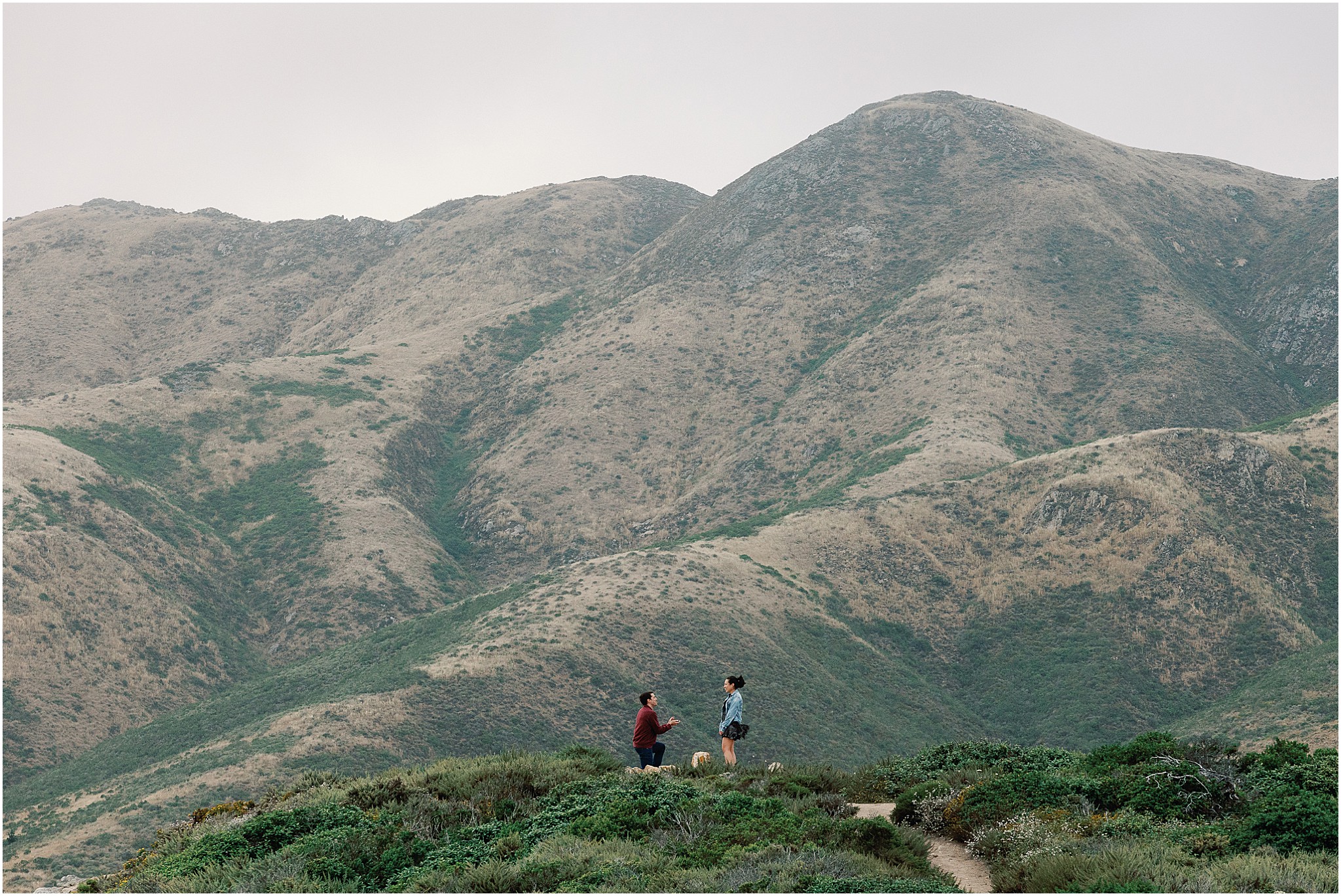 Wide landscape photo capturing Ron proposing to Anna with the Big Sur coastline behind them.