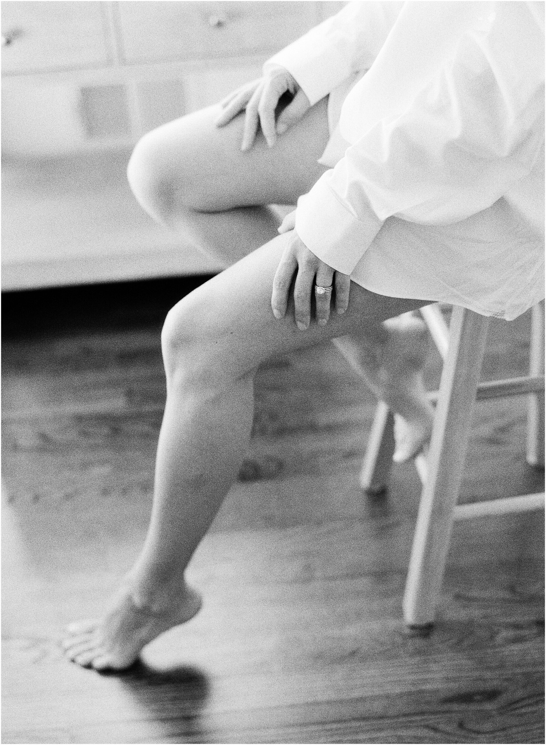 Woman sitting on a chair in an oversized button-down shirt, gently resting her hands on her legs in soft natural light.