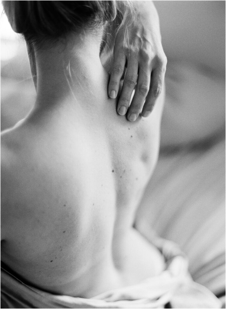 Close-up of a woman’s back and shoulders with her hand resting on her neck in soft, natural light.