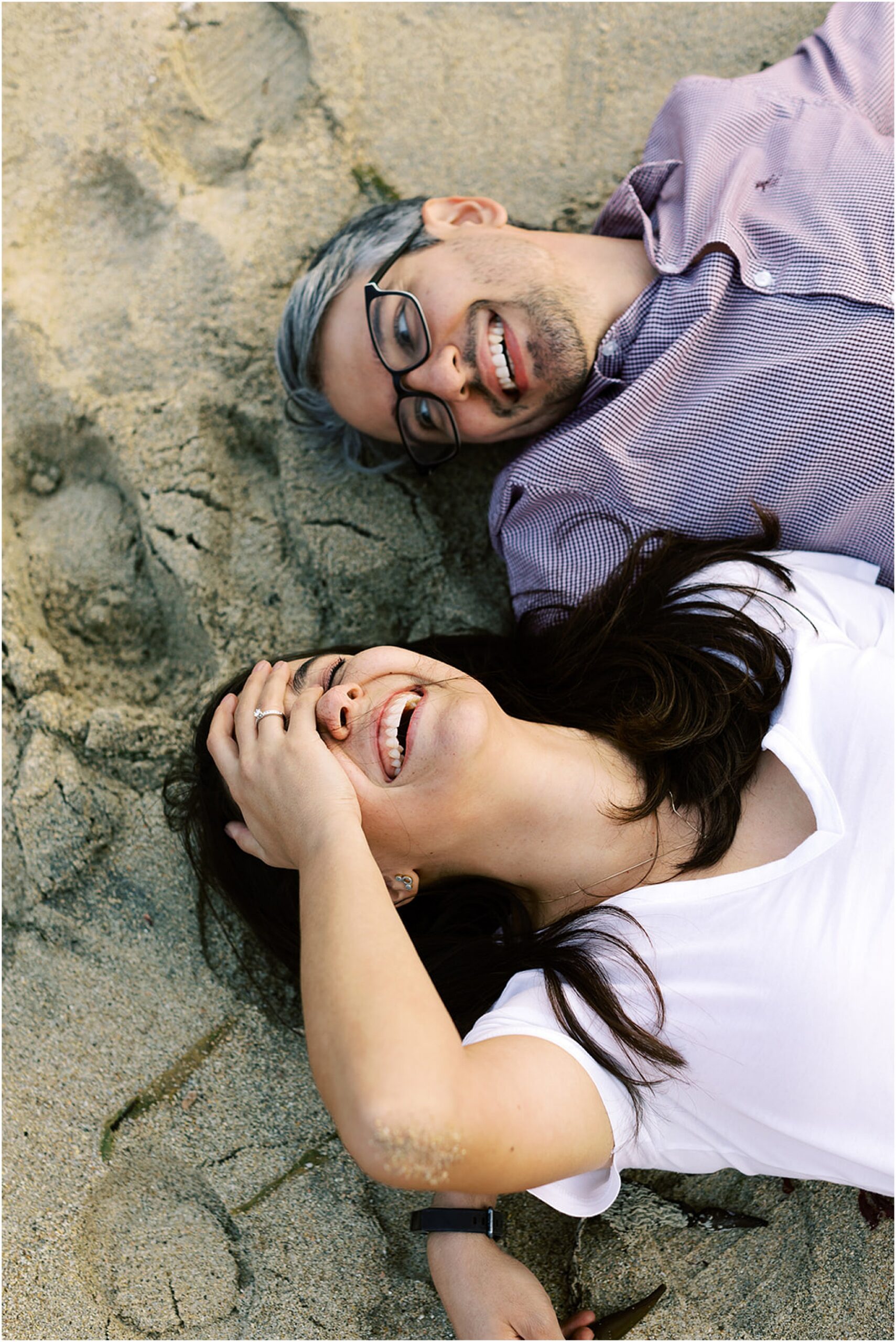 Engaged couple laughing together while relaxing on a sandy California beach.