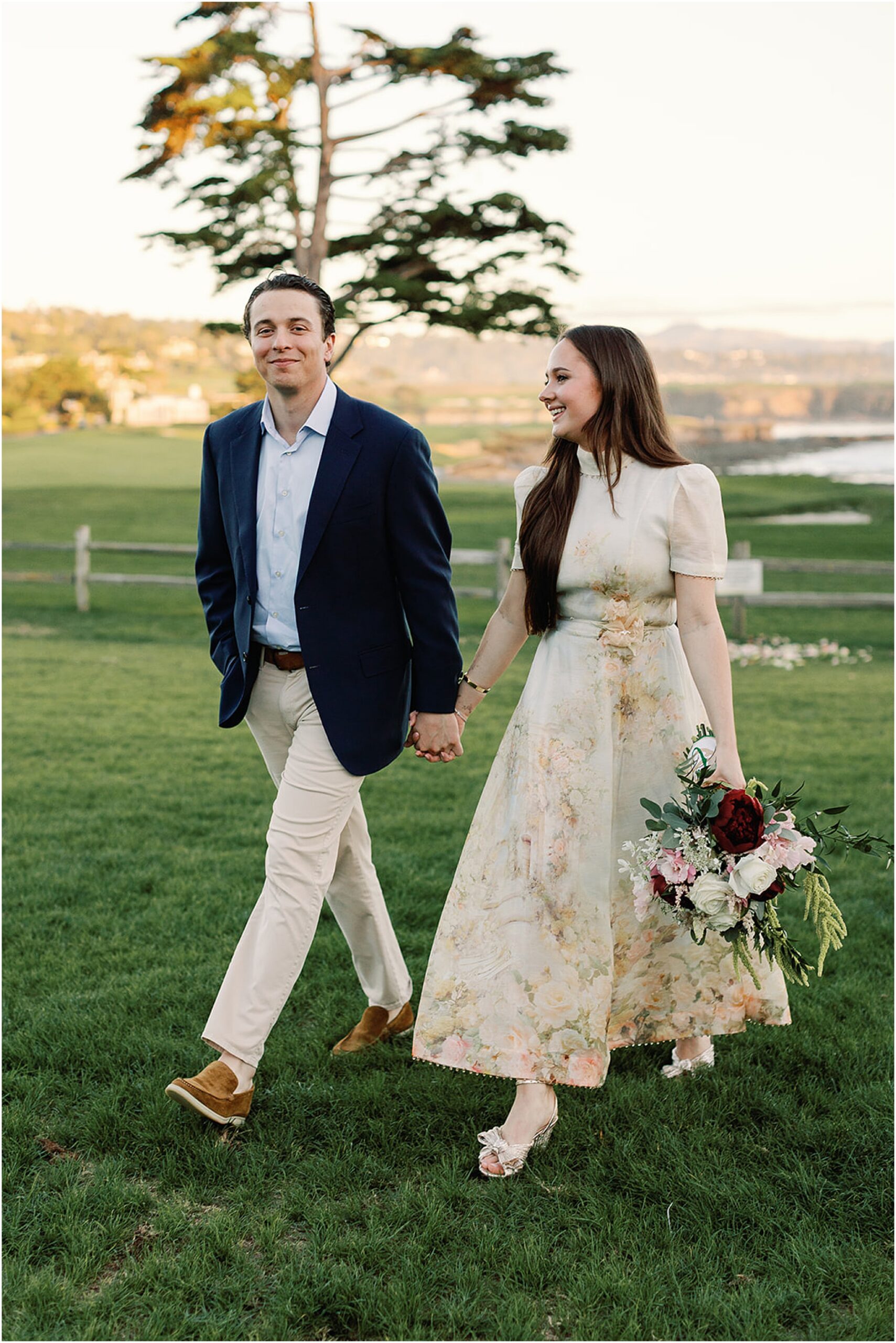 Recently engaged couple walking across the lawn at Pebble Beach carrying flowers.