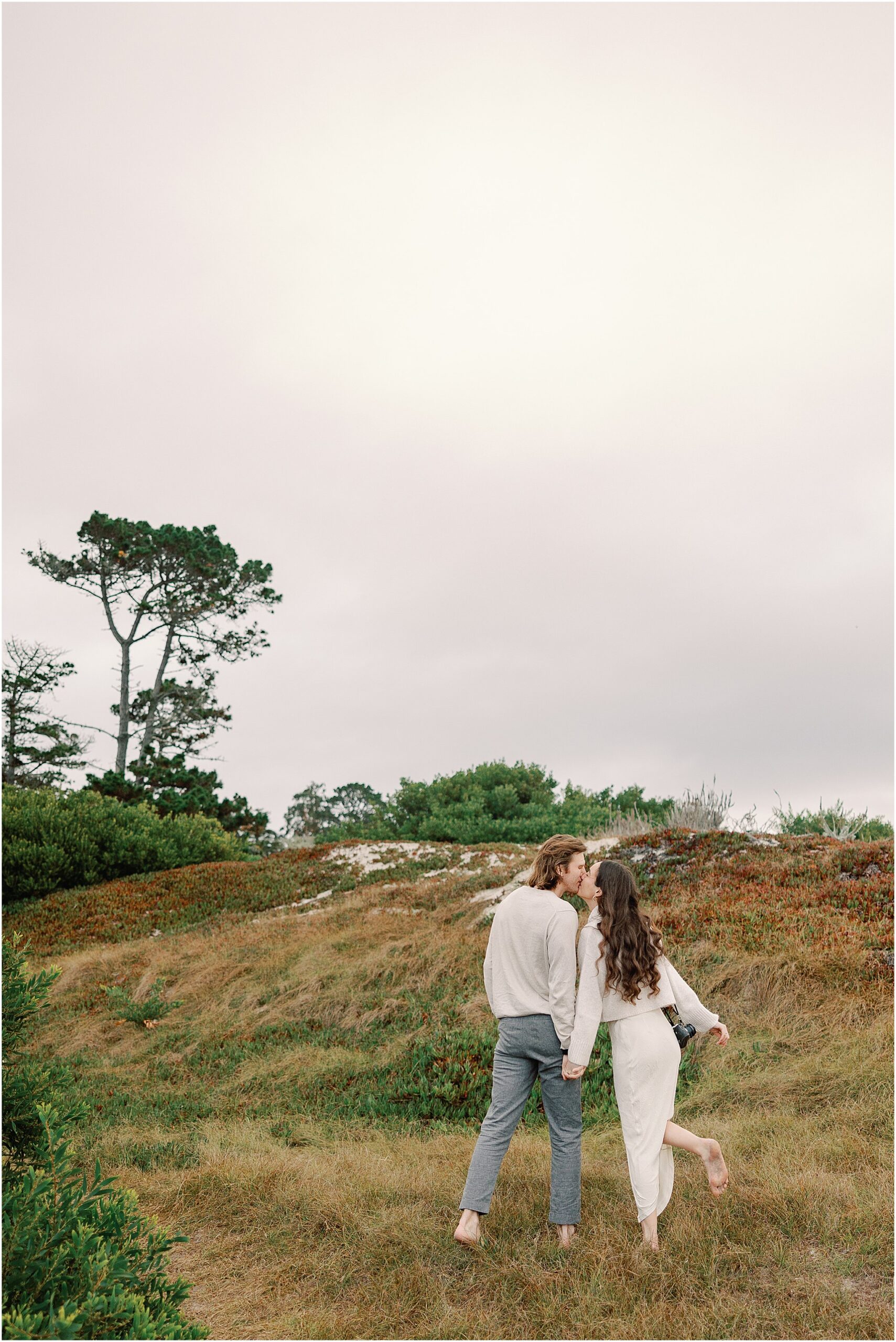 Alex and Connor walking hand in hand through the coastal dunes after their Carmel proposal