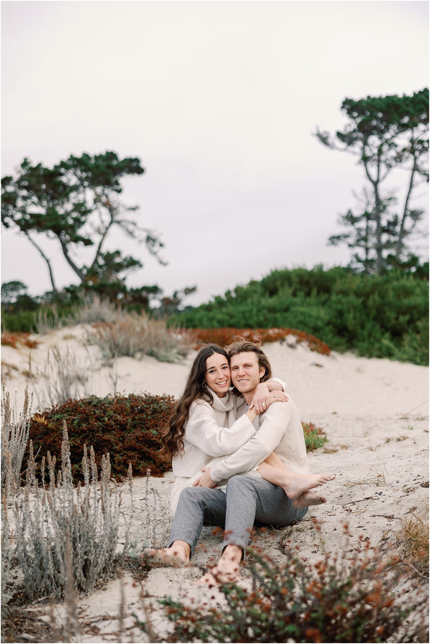 Newly engaged couple Alex and Connor sitting together among the coastal dunes in Carmel