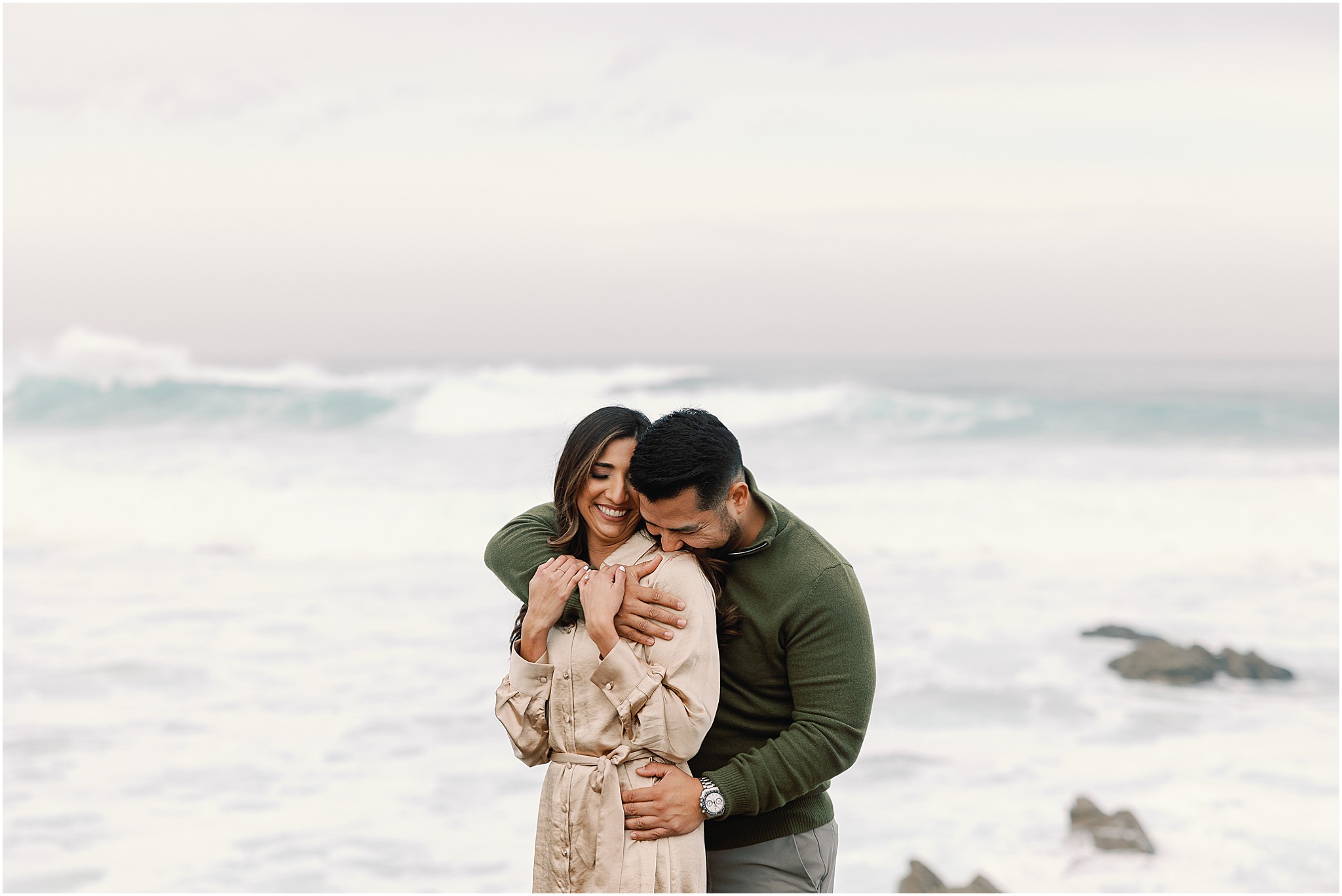 Christine and Carlos laughing together during a playful beach portrait with the ocean behind them